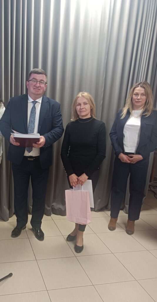 Trzy osoby stoją przed zasłoną; mężczyzna trzyma dokumenty, kobieta w czerni różową torbę. Jasne tło i podłoga z płytek.