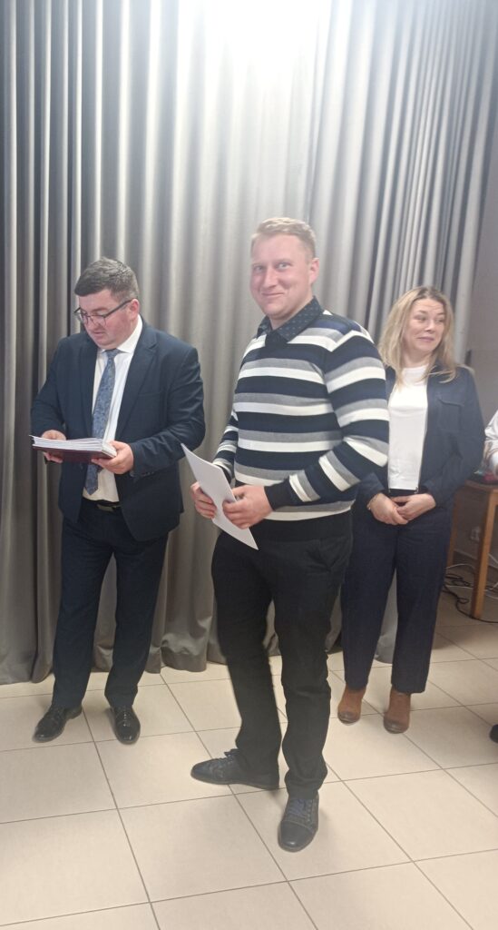 Na zdjęciu widać trzy osoby trzymające dokumenty. Mężczyzna w garniturze i mężczyzna w pasiastym swetrze stoją na tle szarych zasłon. Kobieta w białej bluzce i ciemnej marynarce stoi obok.