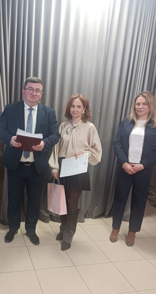 Trzy osoby stoją na tle szarych zasłon. Środkowa kobieta trzyma dokument i różową torbę. Mężczyzna w garniturze trzyma notatki.