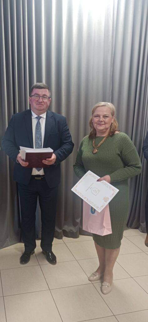 Na zdjęciu jest mężczyzna w garniturze trzymający dokumenty i kobieta w zielonej sukience z dyplomem. Tło zasłania srebrna zasłona. Kobieta trzyma także różową torbę na prezenty.