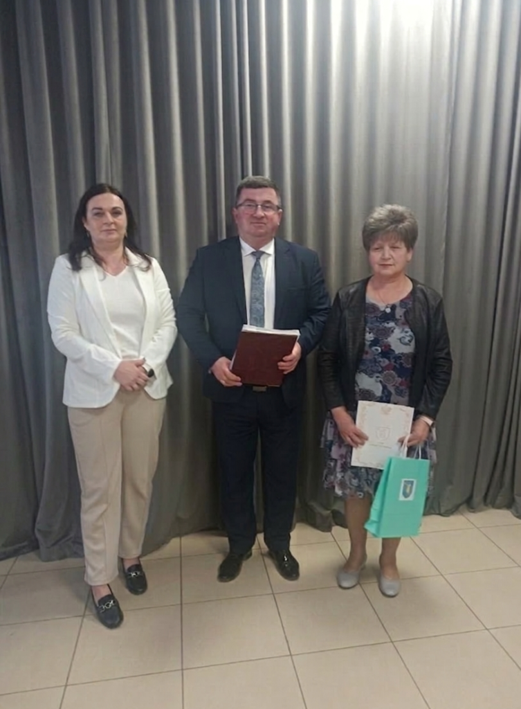 Na zdjęciu widać trzy osoby stojące na tle szarych zasłon. Mężczyzna w garniturze trzyma czerwony folder, kobiety mają białą i czarną odzież. Podłoga jest z jasnych kafelków.