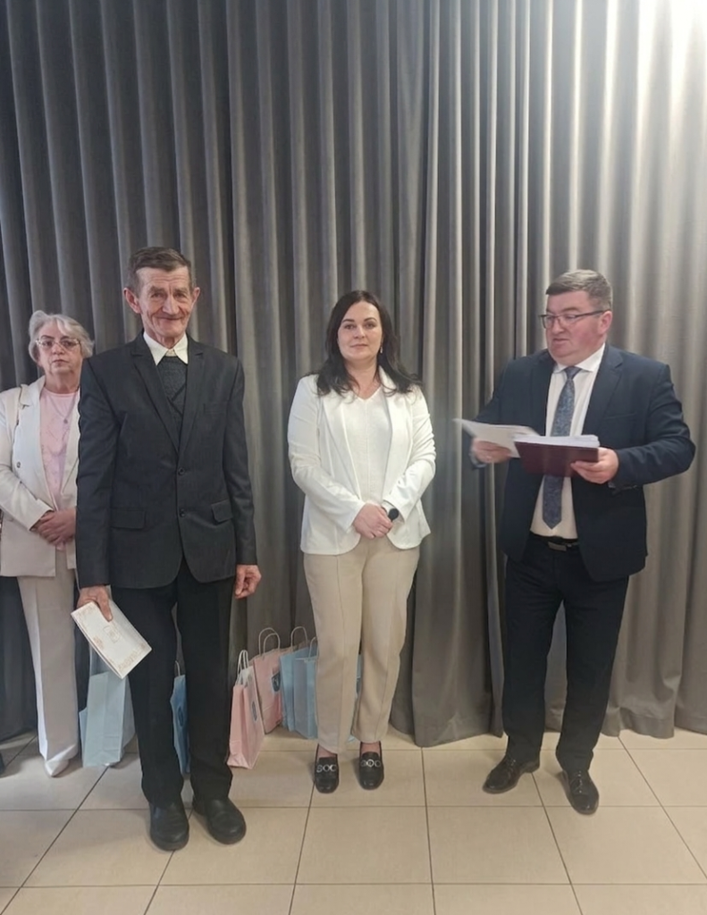 Cztery osoby stoją przed szarymi zasłonami. Trzymają torby i dokumenty. Mężczyzna po prawej czyta coś z kartki.