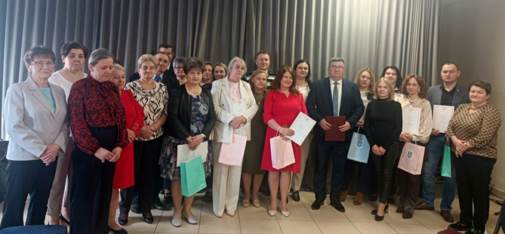 Grupa osób pozuje razem, niektórzy trzymają torby i dokumenty. Kolory ubrań są zróżnicowane, na tle ciemnoszarych zasłon. Elegancki, formalny wygląd sugeruje oficjalne wydarzenie.