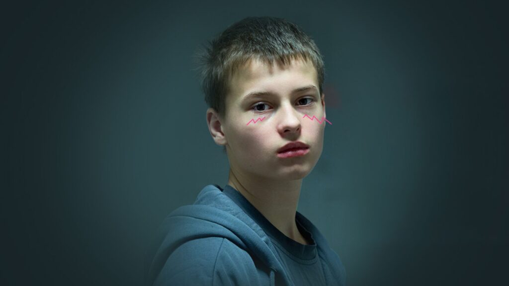 Na ciemnym tle młoda osoba w niebieskiej bluzie patrzy przed siebie. Na policzkach ma różowe, faliste linie. Wyraz twarzy jest poważny.