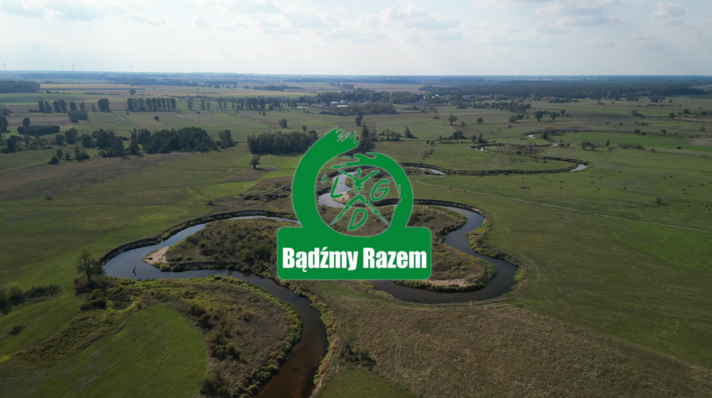 Na zdjęciu widać krajobraz wiejski z rzeką wijącą się przez zielone pola. Na pierwszym planie logo Bądźmy Razem w kolorze zielonym. W tle rozciąga się niebo z delikatnymi chmurami.