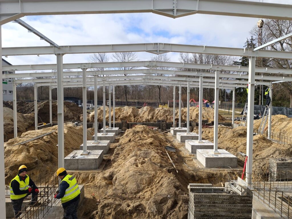 Budowa stalowej konstrukcji z jasnymi belkami i fundamentami na piaskowym podłożu. Pracownicy w żółtych kamizelkach. Tło stanowią drzewa i lekko zachmurzone niebo.