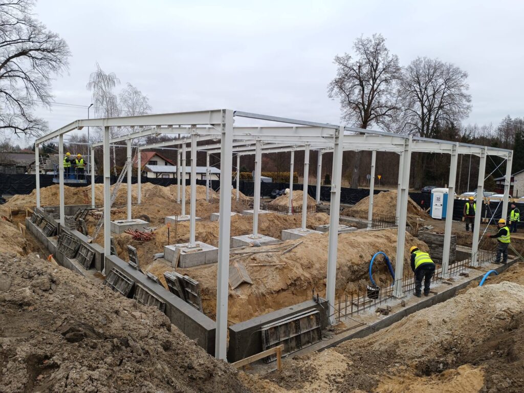 Budowa w toku z białymi stalowymi szkieletami. Pracownicy w żółtych kamizelkach i kaskach. Tło z drzewami i chmurami.