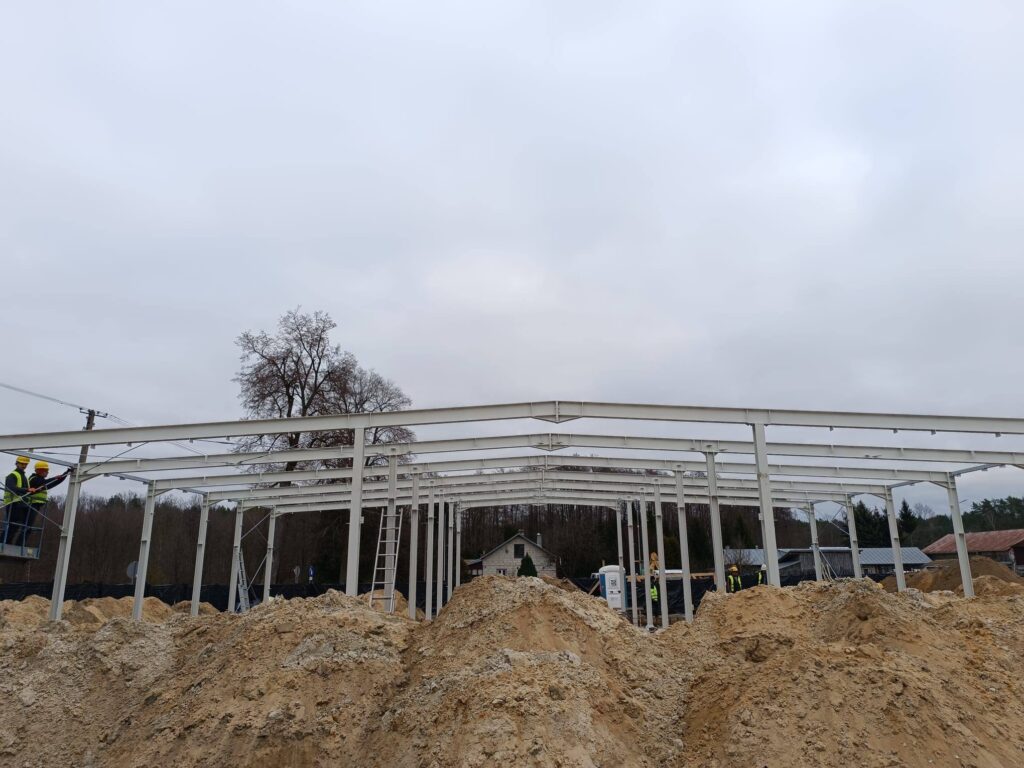 Plac budowy z metalową konstrukcją w tle, robotnicy w żółtych kaskach. Jasne, pochmurne niebo, ziemia w odcieniach beżu.