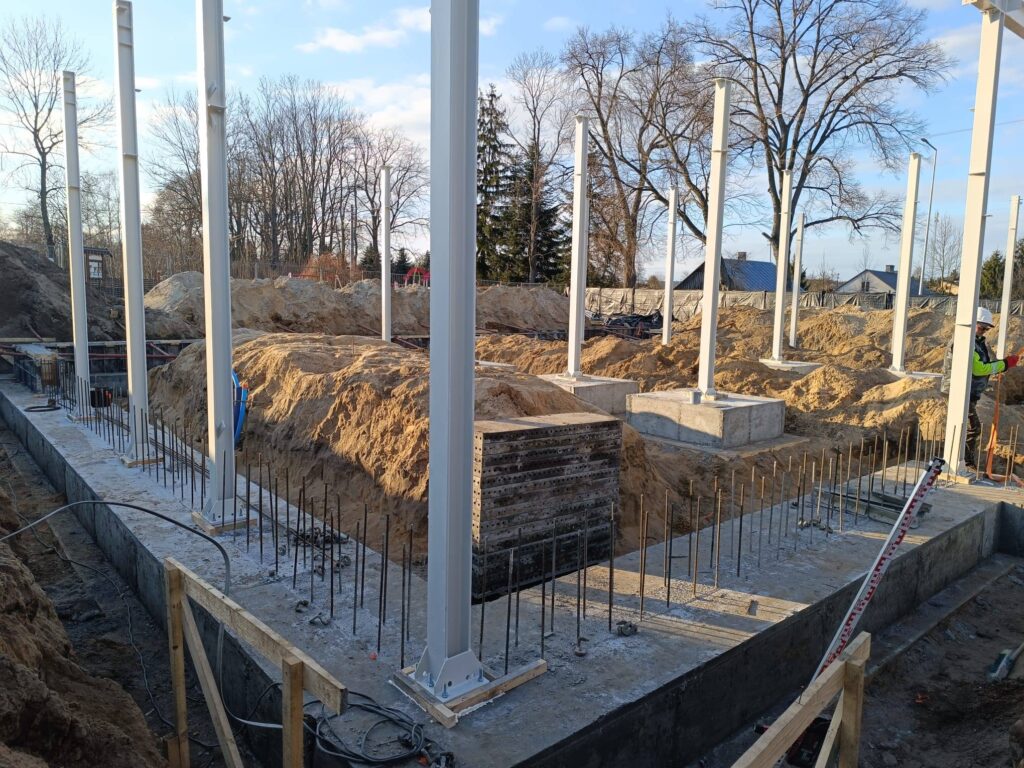 Plac budowy z białymi stalowymi słupami na betonowych fundamentach. W tle drzewa i niebieskie niebo. Mężczyzna w odblaskowej kamizelce stoi obok.