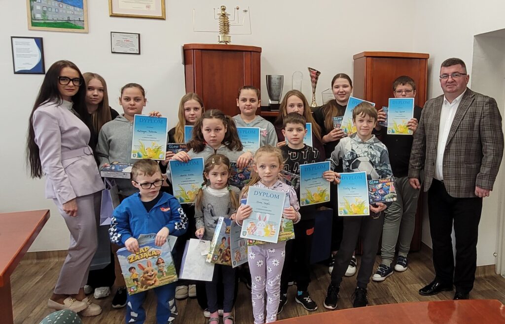 Grupa dzieci w jasnym pomieszczeniu trzyma kolorowe dyplomy, ubrani są różnorodnie. Za nimi stoją dwie osoby dorosłe. Na ścianach obrazy i plakaty, w tle brązowe szafki.