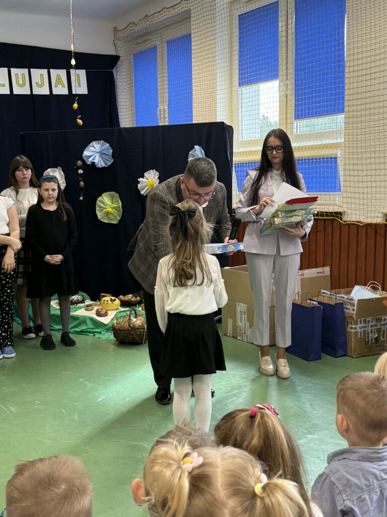 Sala szkolna z dekoracjami na ścianie; mężczyzna wręcza nagrodę dziewczynce. Tło w odcieniach granatu i zieleni.