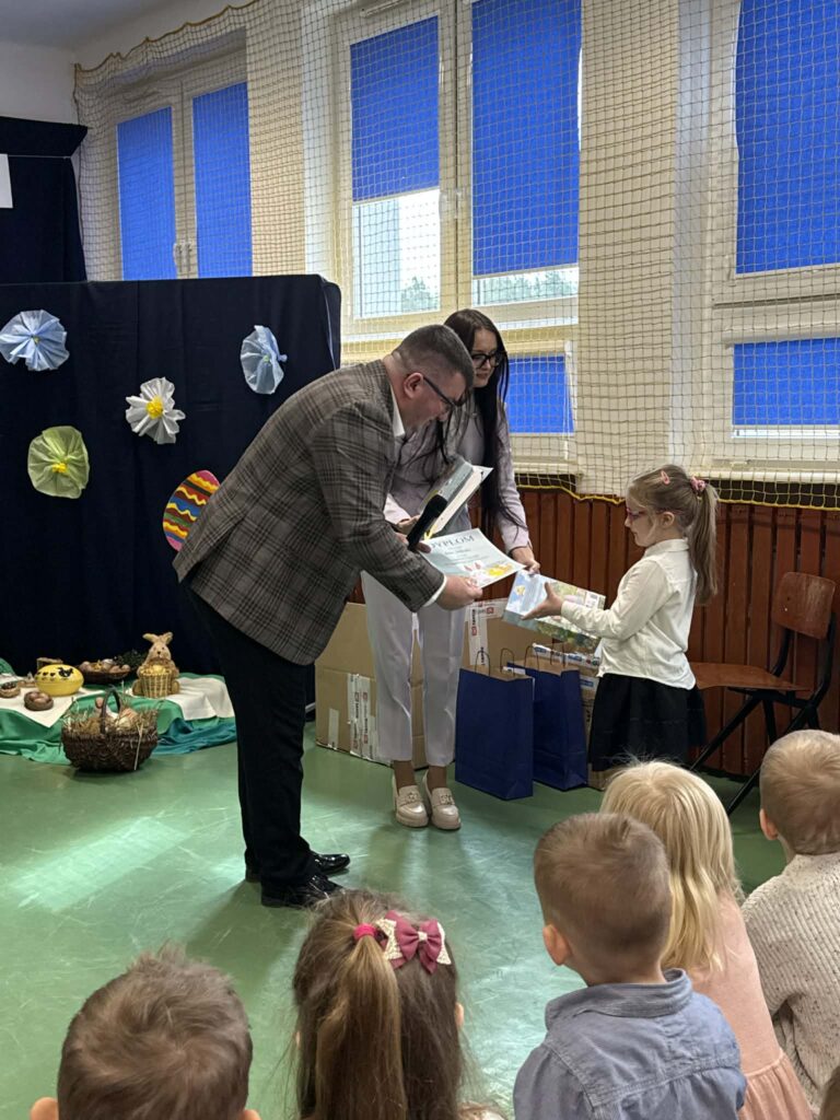 Trzy osoby w sali odbierają nagrodę na tle dekoracji z kolorowymi ozdobami. Podłoga jest zielona, a w tle niebieskie zasłony. Dzieci siedzą na pierwszym planie.