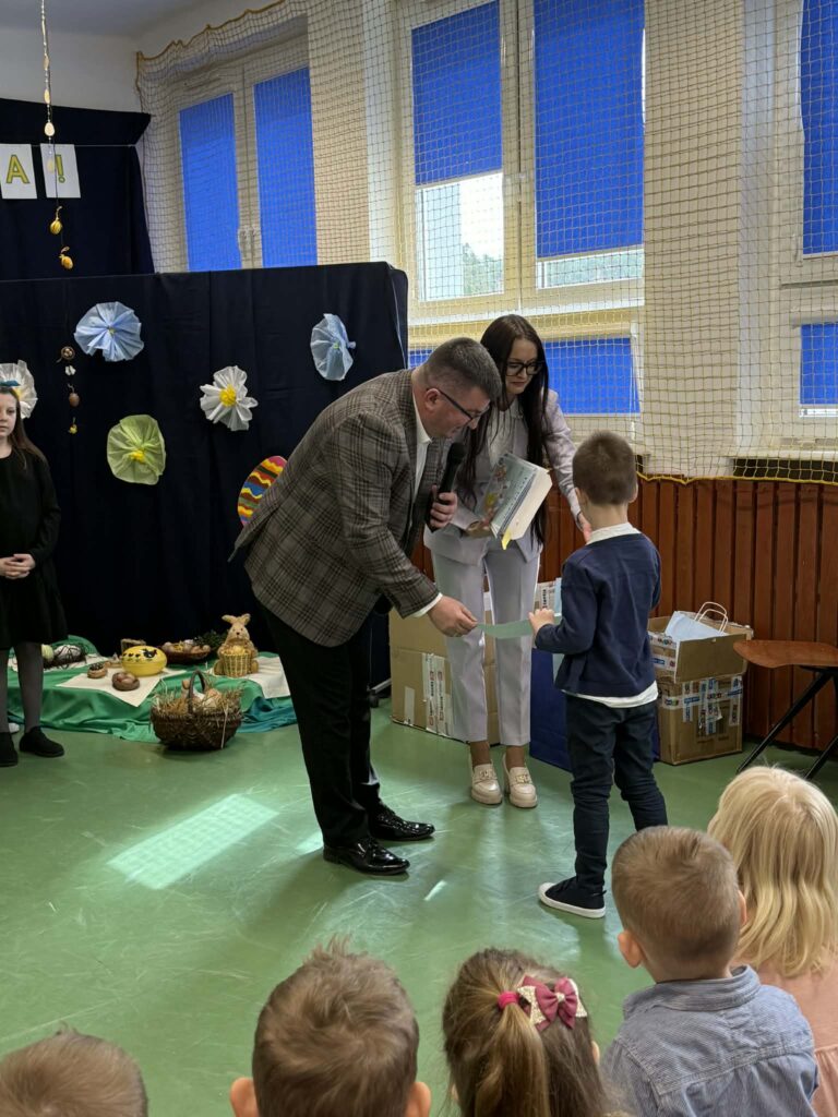 W sali z zieloną podłogą dorosły wręcza dziecku nagrodę. Tło ozdobione jest kwiatami w odcieniach niebieskiego i żółtego. W tle widać okna z niebieskimi zasłonami.