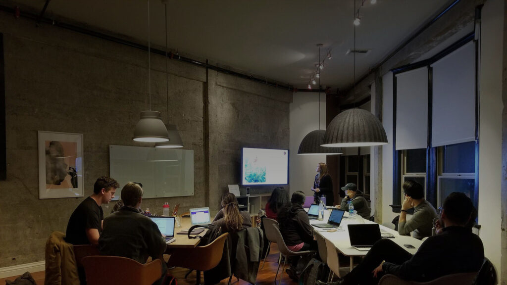 Grupa osób siedzi przy dużym stole z laptopami w sali konferencyjnej. Na ścianie wisi duży ekran, a tło jest w szarym kolorze. Pomieszczenie oświetlone jest wiszącymi lampami.