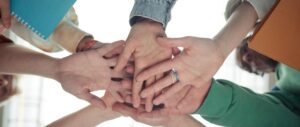 Kilka osób łączy dłonie w stosie, symbolizując współpracę. Różnorodne kolory rękawów dodają barw. Tło jasne i rozmyte.