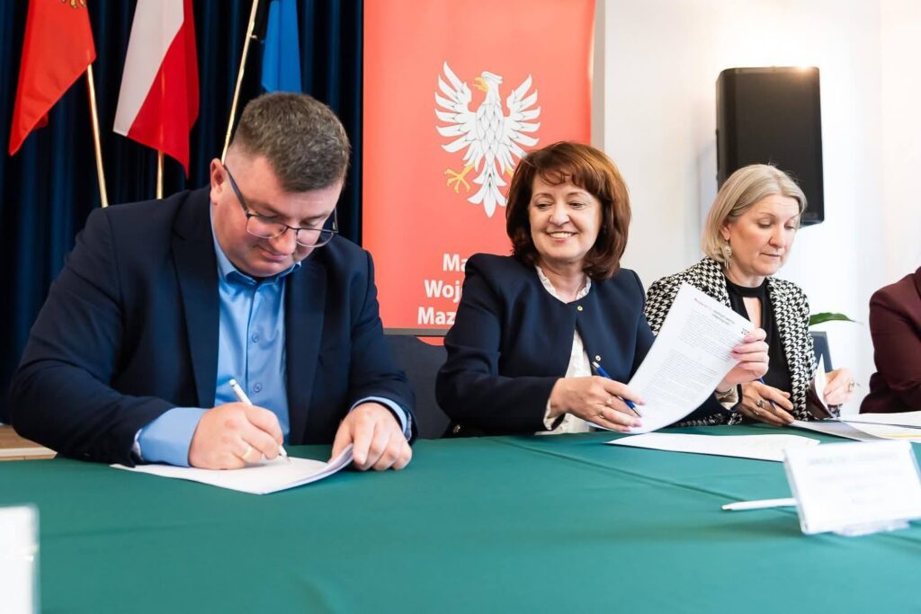 Trzy osoby siedzą za zielonym stołem, podpisują dokumenty. W tle widać czerwony baner z orłem. Kolory flag i ubrania są wyraziste.