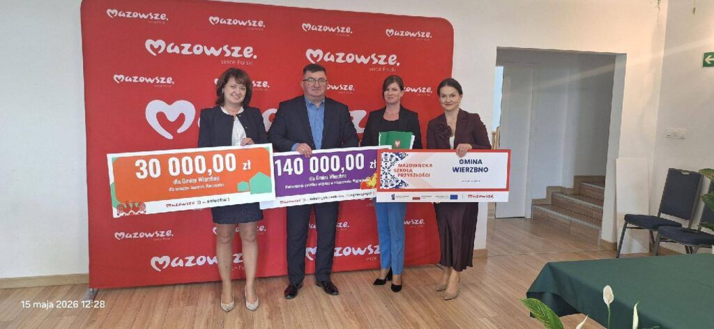 Cztery osoby trzymają czeki na tle czerwonego baneru z napisem Mazowsze. Kolory czeków to pomarańczowy, fioletowy i biały. W tle jasna ściana i podłoga.