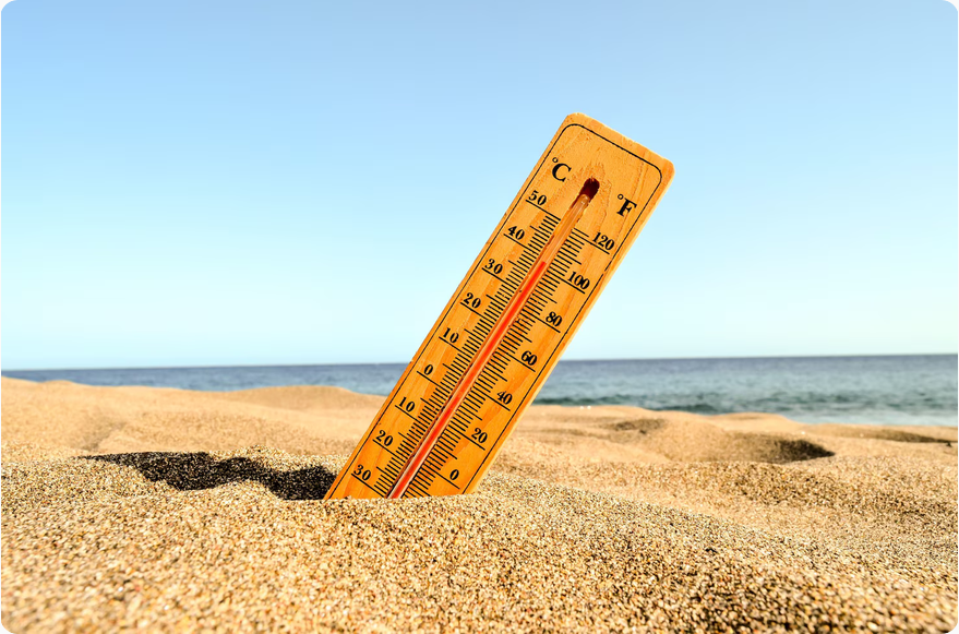 Drewniany termometr wbity w piasek na plaży. Jest słoneczny dzień z błękitnym niebem i morzem w tle. Temperatury oznaczone są w stopniach Celsjusza i Fahrenheita.