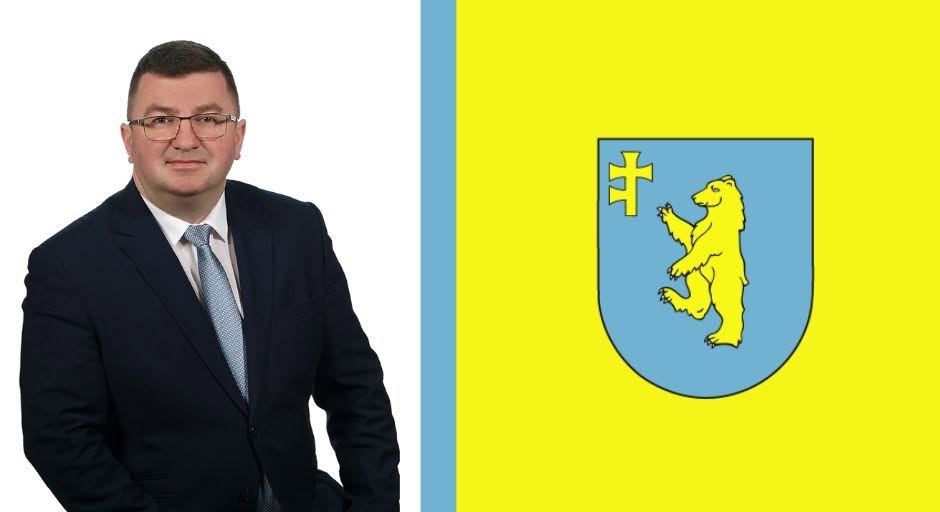 Na zdjęciu widać mężczyznę w garniturze na białym tle. Obok znajduje się flaga z żółtym niedźwiedziem na niebieskim tle. Całość utrzymana w prostym, kontrastowym układzie.