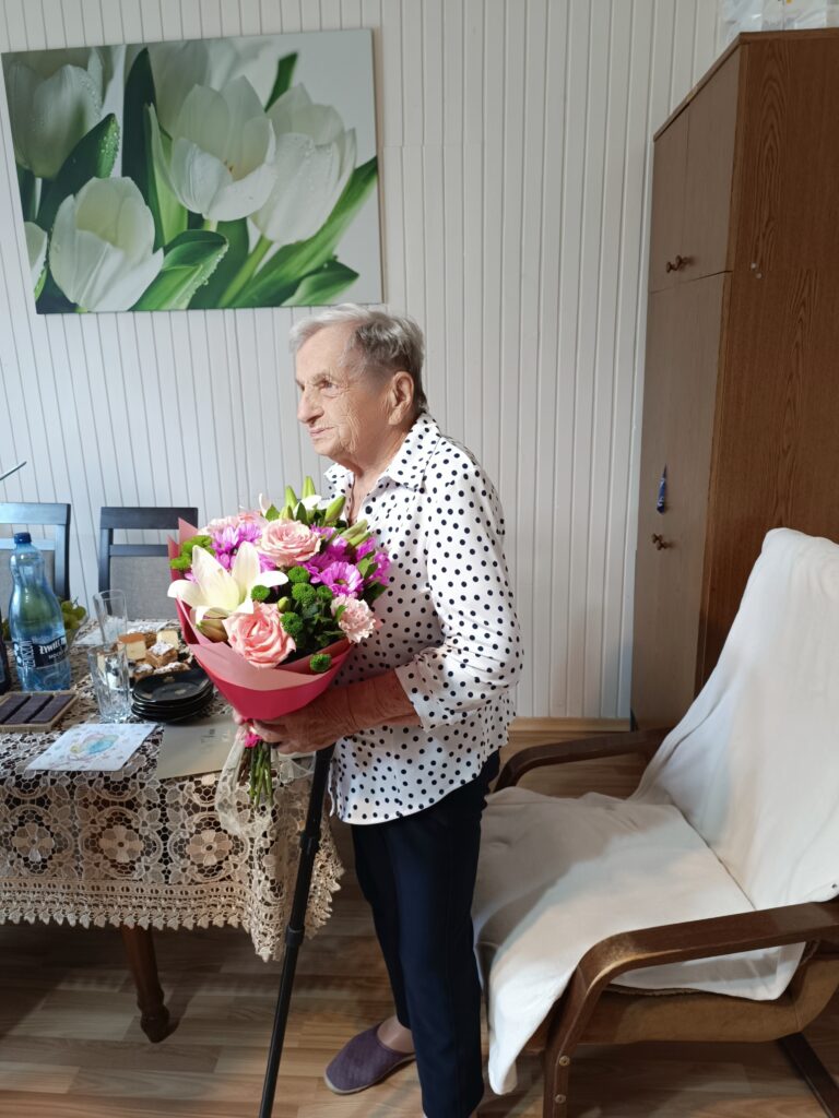 Starsza kobieta w białej bluzce w kropki trzyma bukiet kwiatów. W tle jasna ściana i obraz z tulipanami. Obok znajduje się stół z koronkowym obrusem.
