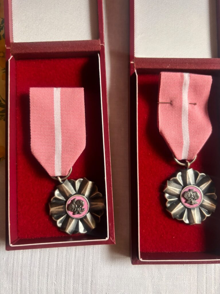 Dwóch różowych medali leżących w czerwonych pudełkach na białym tle. Każdy medal ma metalowe kwiaty w środku.