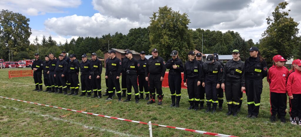 Grupa strażaków w czarnych mundurach stoi na trawie, w tle las i chmury na niebie. Obok dzieci w czerwonych strojach. Po lewej stronie czerwone samochody strażackie.
