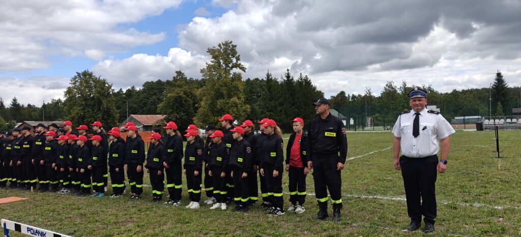 Grupa ludzi w strojach strażackich i czerwonych czapkach stoi na trawniku. W tle są drzewa i budynki pod pochmurnym niebem. Mężczyzna w białej koszuli stoi po prawej stronie.