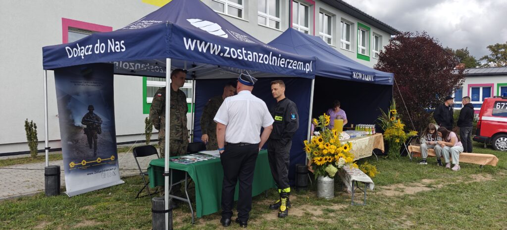 Namioty promocyjne z hasłami rekrutacyjnymi na trawie przed budynkiem, w tle stoły z napojami i słoneczniki. Na pierwszym planie mężczyźni w stroju wojskowym i strażackim, rozmawiający obok stolika. Obok