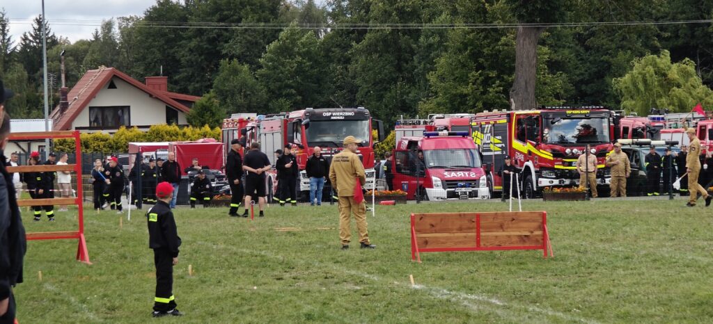 Strażacy stoją na zielonej trawie przed czerwonymi wozami strażackimi. Na tle drzewa i budynku widać ludzi w mundurach. W centrum drewniana przeszkoda.