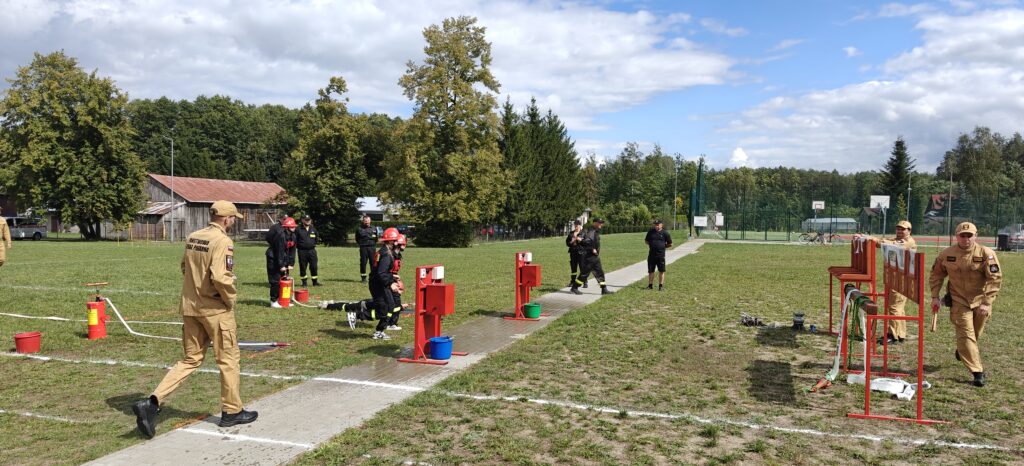 Grupa strażaków w beżowych i czarnych uniformach na zielonym boisku, wykonuje ćwiczenia z gaśnicami. W tle stoi drewniany budynek oraz drzewa. Jasne niebo częściowo pokryte chmurami.