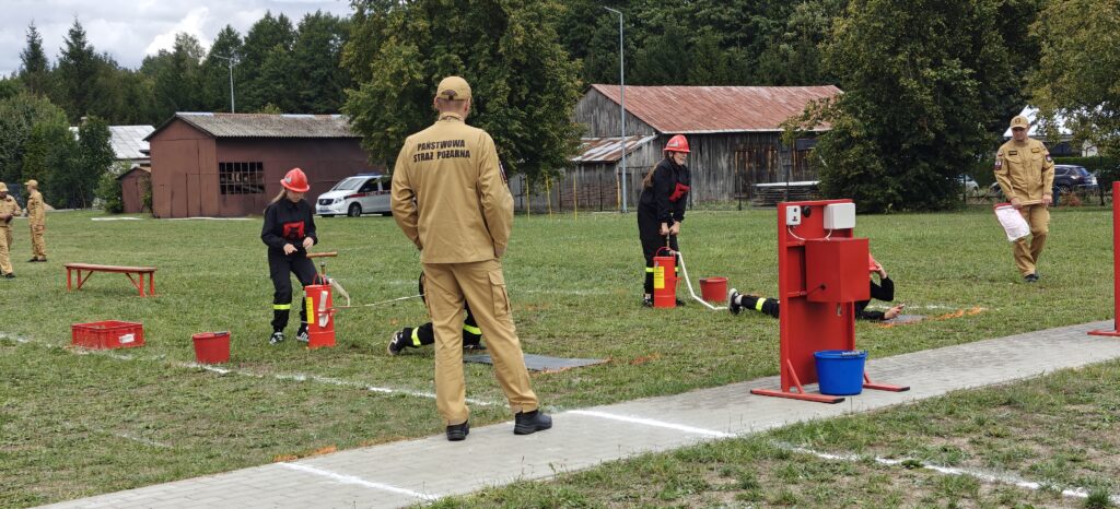 Osoby w strojach strażackich przeprowadzają ćwiczenia na trawie. Tło stanowią drewniane budynki i drzewa. Widoczne są czerwone akcenty sprzętu.