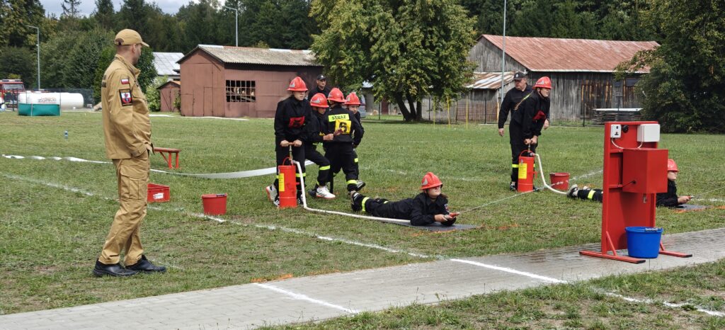 Na trawie młodzież w strojach strażackich z czerwonymi hełmami uczestniczy w ćwiczeniu z wężem. Z tyłu budynki i drzewa. Na pierwszym planie czerwona konstrukcja i wiadro.