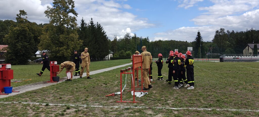 Grupa strażaków w mundurach ćwiczy na zielonym polu z czerwonymi hydrantami. Na niebie widać chmury i błękitne niebo. Drzewa w tle tworzą zielony krajobraz.