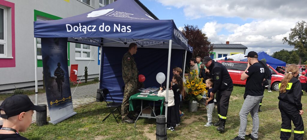 Namiot wojskowy z napisem Dołącz do Nas, w tle kolorowy budynek. Żołnierz rozmawia z dziećmi. Obok czerwony wóz strażacki.