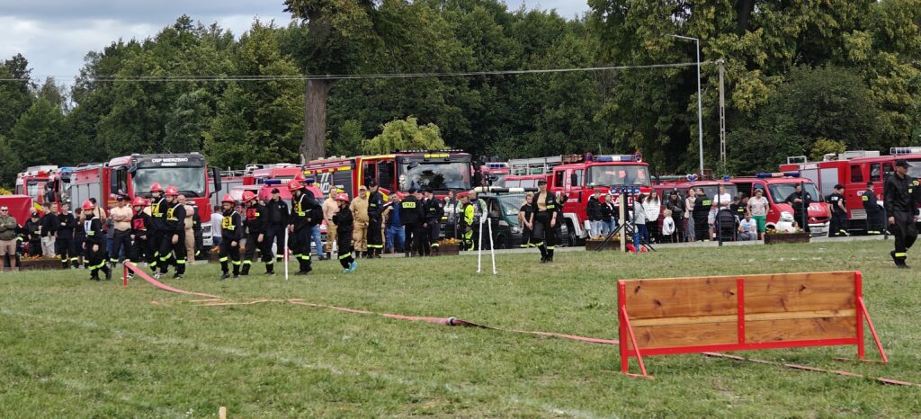 Na zdjęciu widać grupę strażaków w hełmach na zielonej polanie. W tle stoją czerwone wozy strażackie, otoczone przez ludzi. Krajobraz uzupełnia zielone drzewo.