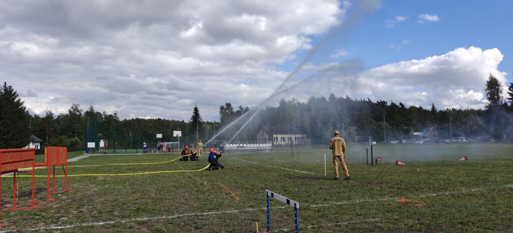 Strażacy w czerwonych kaskach kierują węże wodne na trawiaste pole. Tło to las i niebo z białymi chmurami. Po bokach widoczne są elementy sportowe.