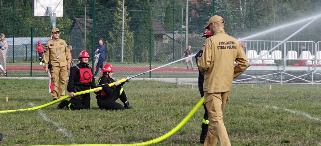 Grupa strażaków w beżowych i czarnych mundurach trzyma wąż strażacki na trawiastej nawierzchni. W tle znajduje się ogrodzenie i ławki. Dzień jest słoneczny, a scena odbywa się na świeżym powietrzu.