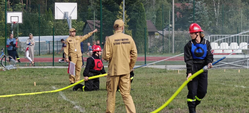 Na zdjęciu widać strażaków w mundurach na zielonej trawie, trzymających żółte węże. W tle znajduje się metalowe ogrodzenie oraz boisko. Osoba w beżowym mundurze stoi przodem do kamery.