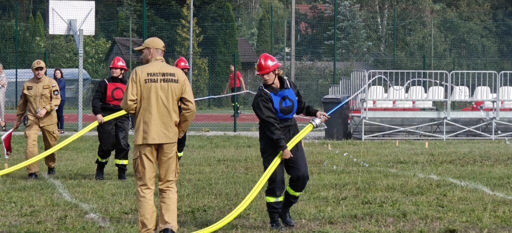 Na zdjęciu kilku strażaków na boisku. Noszą czerwone hełmy i trzymają żółty wąż. W tle widać trybuny i zieleń.