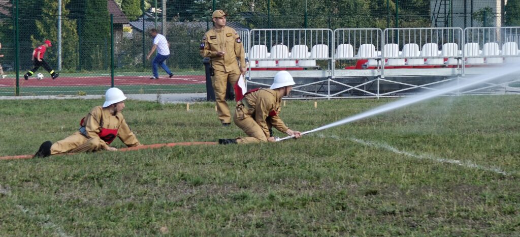 Strażacy w beżowych mundurach trenują z wężem strażackim. Tło to zielona trawa i widownia z białymi krzesłami. Przy boisku widać biegające osoby.