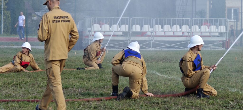 Strażacy w beżowych mundurach ćwiczą z wężami na zielonej trawie. Białe kaski i niebieskie oznaczenia widoczne. Tło z metalowymi barierkami i siedzeniami.