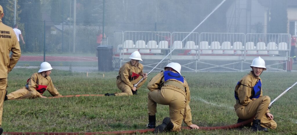 Na trawie są czterej strażacy w jasnobrązowych uniformach i białych kaskach. Trzymają wąż strażacki, a w tle widać trybuny. Delikatna mgła unosi się nad polem.