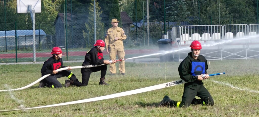 Trzech strażaków z czerwonymi kaskami trzyma węże gaśnicze. Na trawie, w tle siatka i drzewa. Jeden obserwator w mundurze.