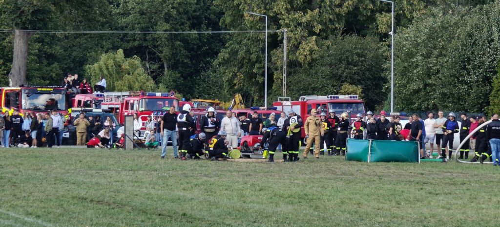 Strażacy w hełmach uczestniczą w wydarzeniu na trawniku, w tle czerwone wozy strażackie i zielone drzewa.
