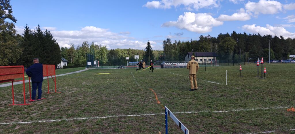 Osoby w mundurach strażackich biorą udział w ćwiczeniach na zielonym boisku. Widać pomarańczowe pachołki i stojące przeszkody. W tle las i niebieskie niebo z chmurami.