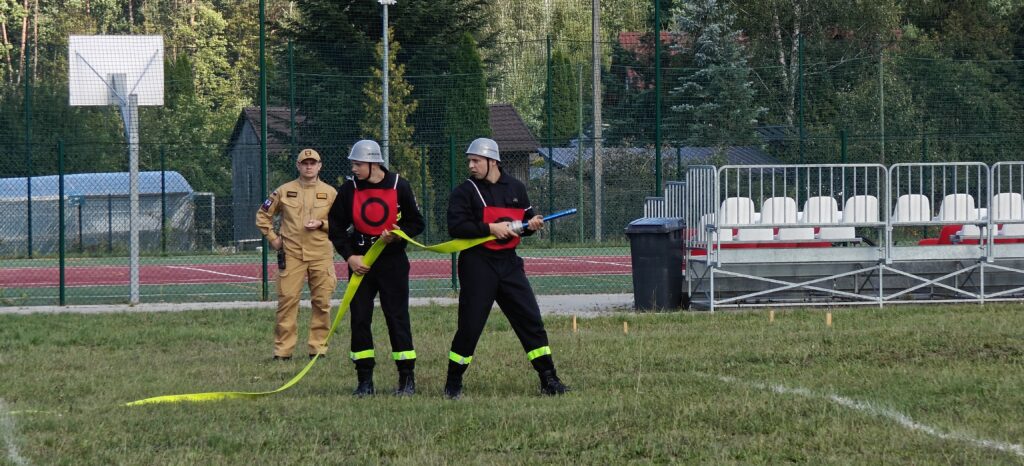 Trzech strażaków na trawie z wężami strażackimi. Dwóch w czarnych uniformach, jeden w beżowym. Tło to zielone drzewka i ogrodzenie.