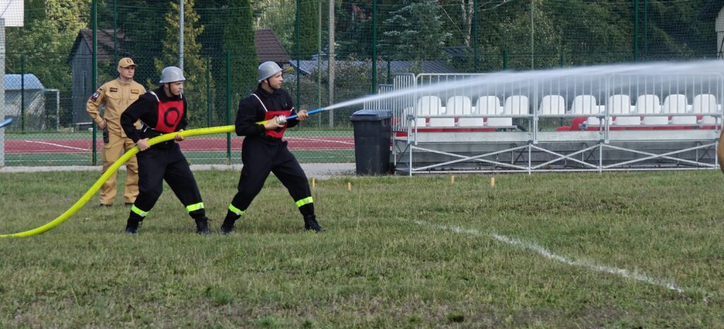 Na zdjęciu dwaj strażacy trzymają wąż i rozpylają wodę. Noszą czarne mundury z odblaskowymi elementami. W tle widać trybuny i boisko sportowe.
