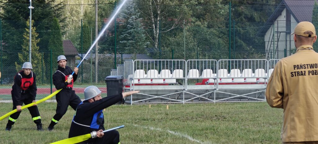 Na obrazie widać strażaków w hełmach podczas ćwiczeń z wężem strażackim. Tło jest leśne, a z boku widać trybuny. Mundury mają czerwone i czarne elementy.