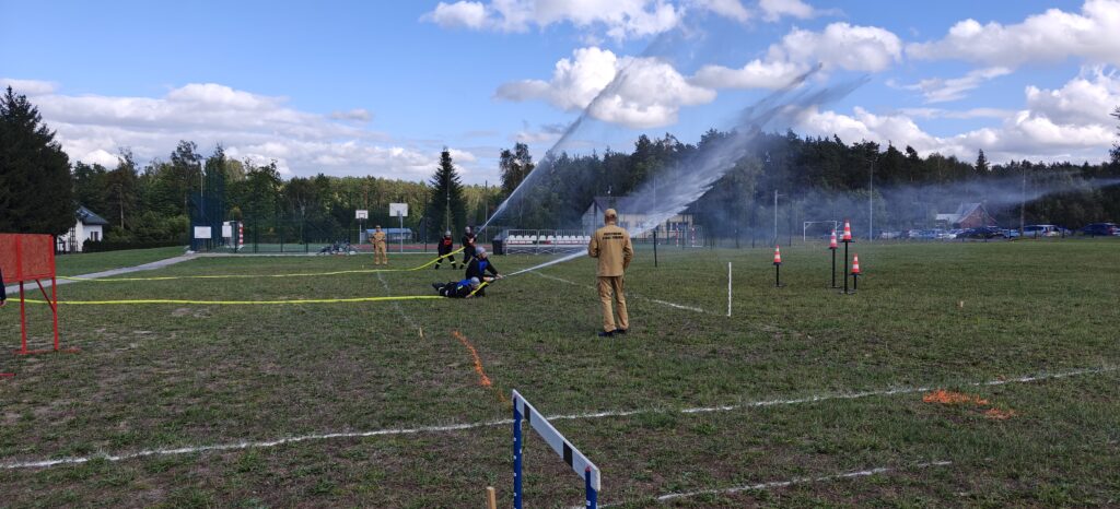 Na boisku zielonej trawy strażacy w mundurach manewrują wężami strażackimi. Niebieskie i białe niebo w tle z chmurami. W oddali drzewa i siatki treningowe.