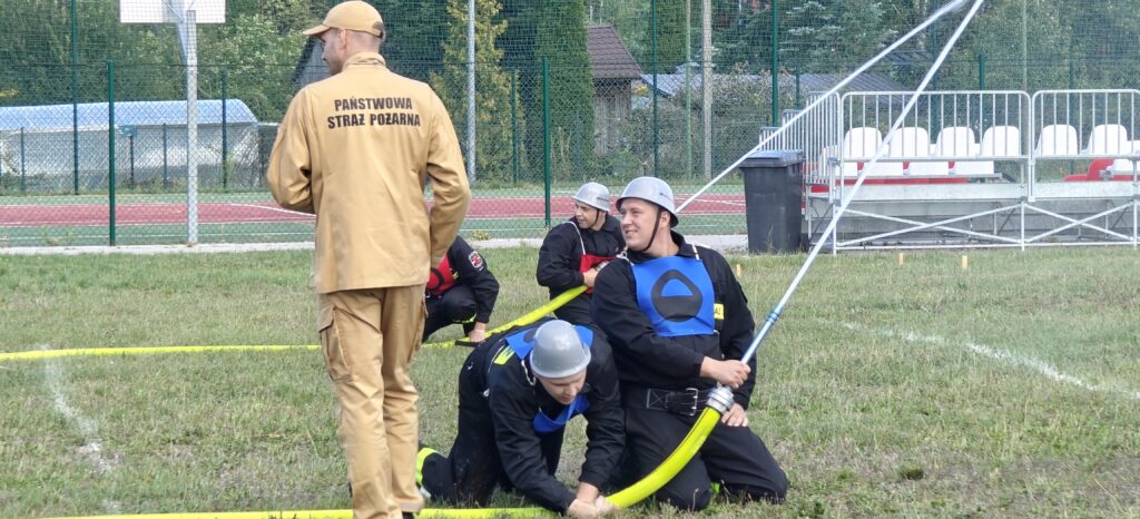 Strażacy w czarnych uniformach ćwiczą na zielonym trawniku. Jeden strażak w beżowym ubraniu stoi z tyłu. W tle widać czerwony tor i białe trybuny.